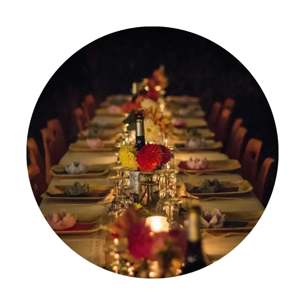 A long, candlelit banquet table set for a shared meal, with plates, folded napkins, glassware, flowers, and bottles arranged down the centre, creating a warm and inviting atmosphere.